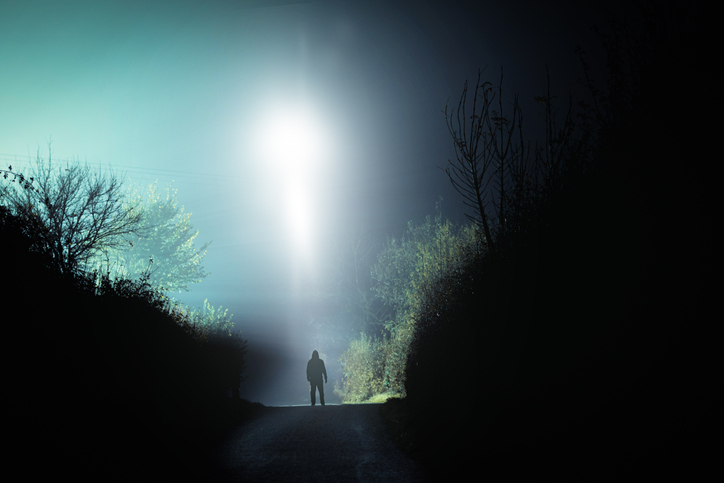 A mysterious scary UFO above a hooded figure. On a spooky country road. On a foggy winters night.