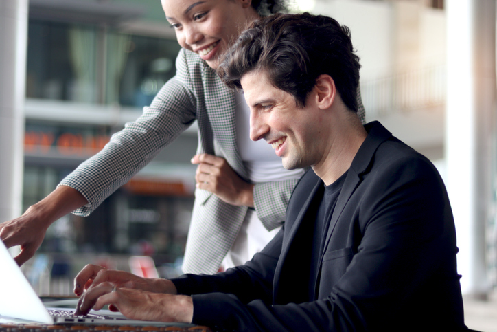 Happy businessman working on laptop on office desk while discussing with his female colleague coworker. Business officers working together at workplace. Successful team of businesspeople at work.