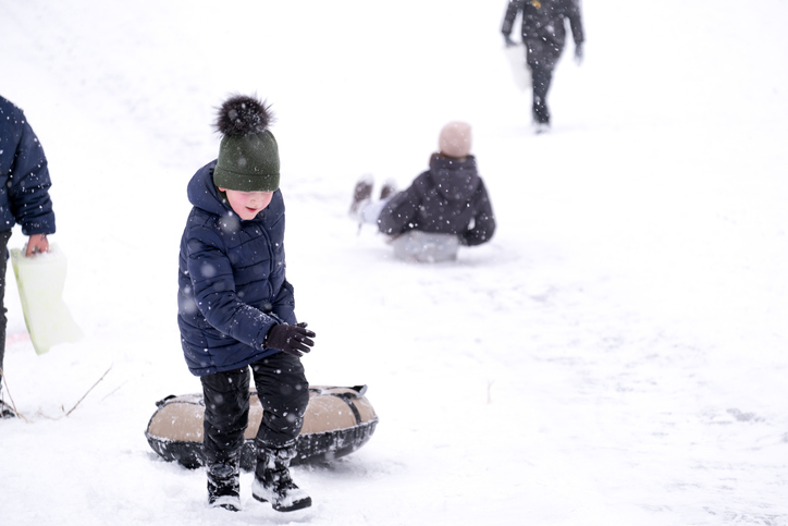A little pretty boy in a dark overalls slides down an ice slide, winter children's games, a child in the park in winter. The boy climbs the slide with tubing