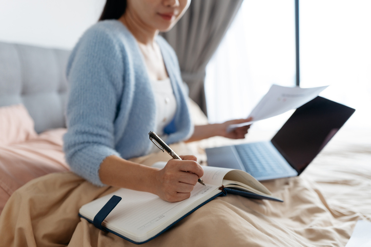Close up of Asian woman writing on notebook and using laptop on bed
