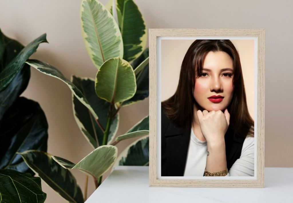 A framed portrait of a young woman with long dark hair and red lipstick, set against a background of lush green leaves.