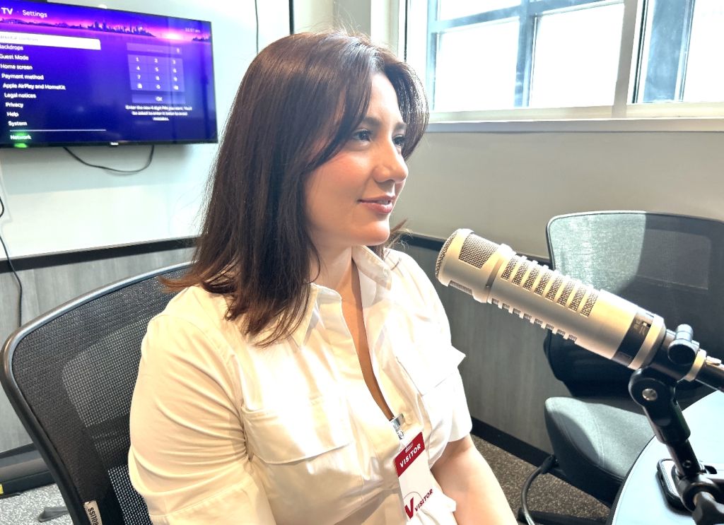 A woman wearing a white shirt and holding a microphone in a studio setting with a TV screen displaying settings in the background.