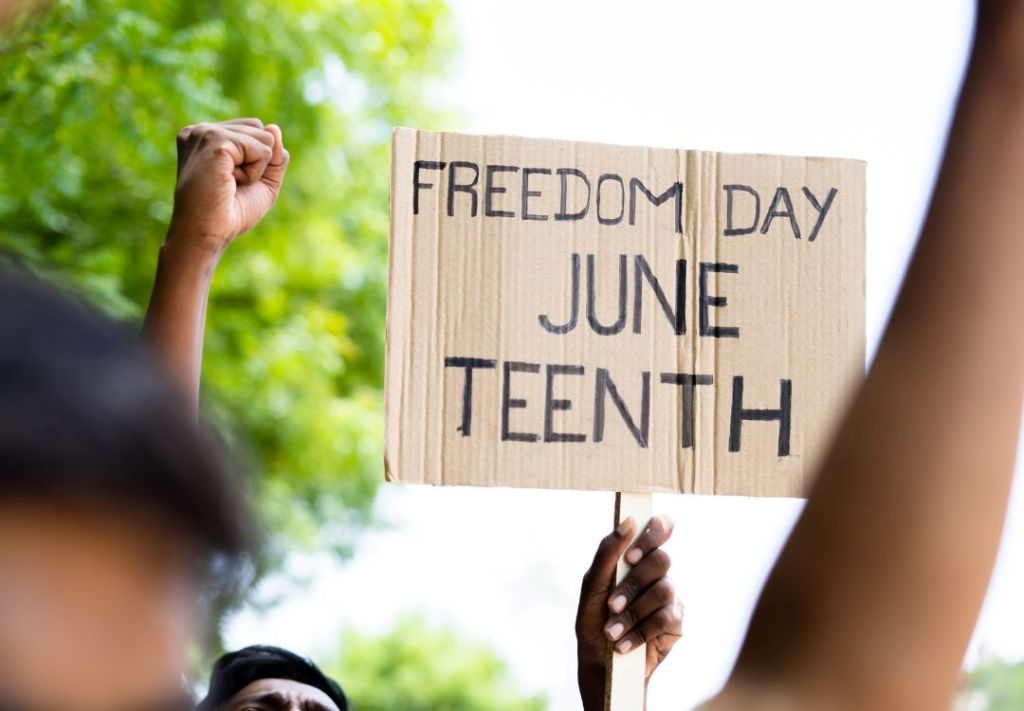 A person holding a cardboard sign that reads "FREEDOM DAY JUNE TEENTH" with a raised fist.