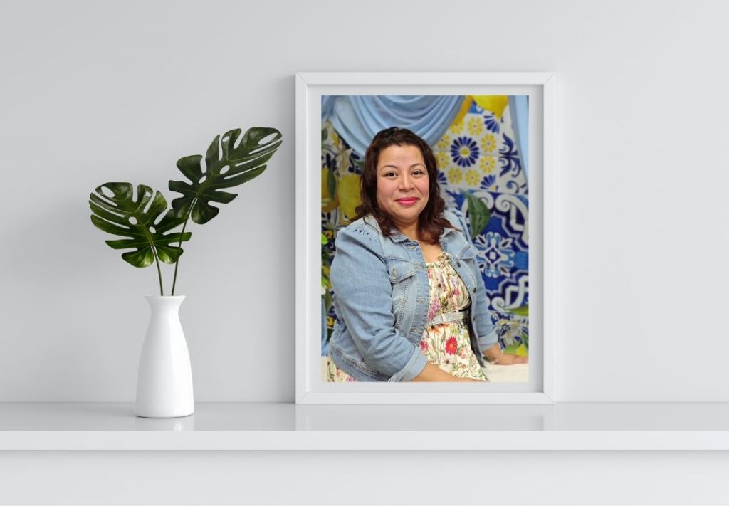 A smiling woman wearing a floral top and denim jacket stands in front of a colorful patterned backdrop.