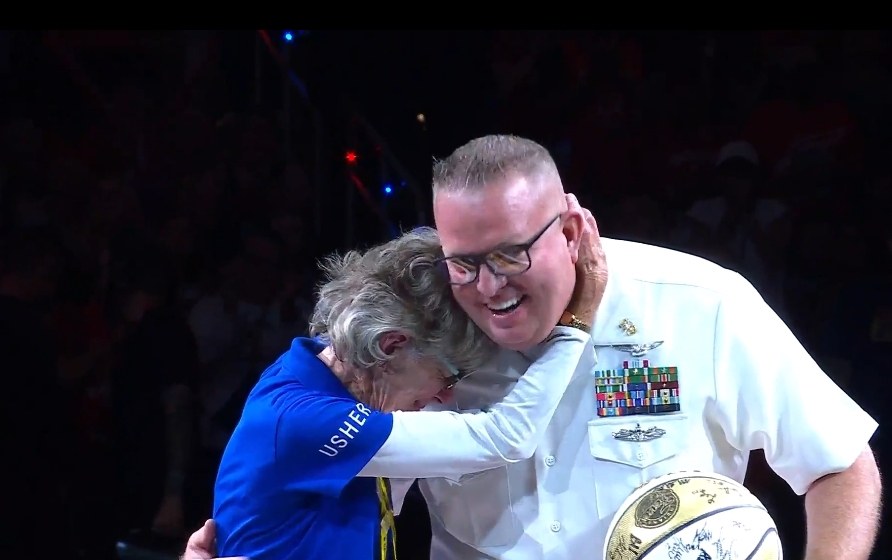 A man in military uniform embracing an elderly woman wearing a "USHER" jacket, both with joyful expressions.