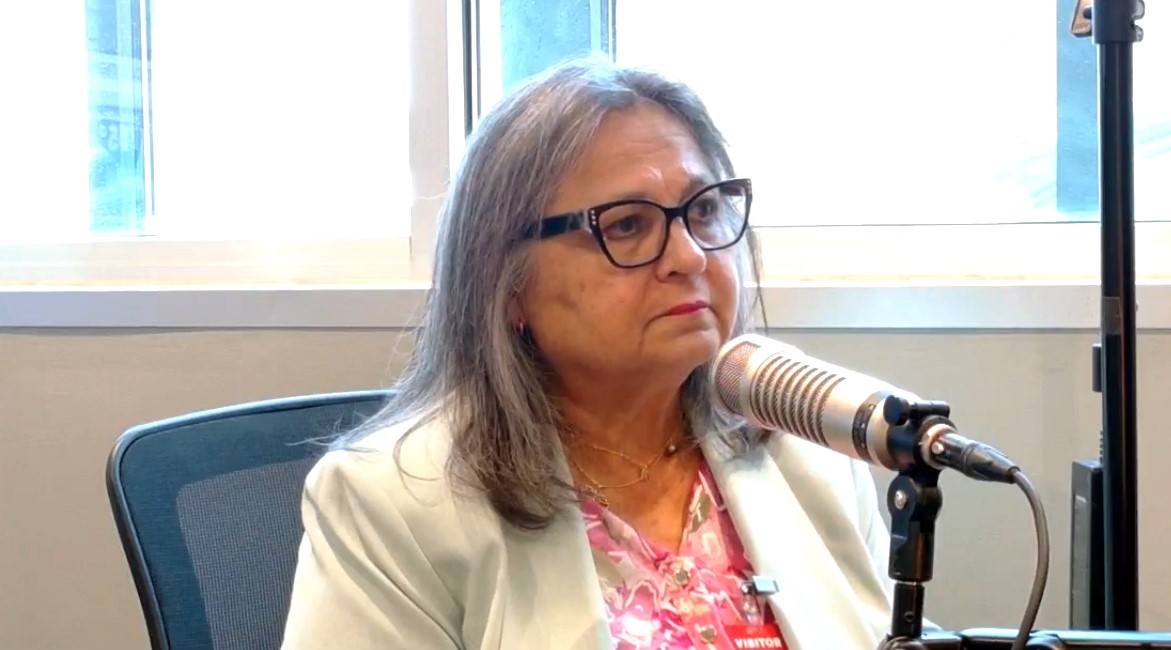 A woman with glasses and long gray hair speaking into a microphone at a desk in front of a window.