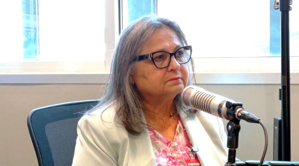 A woman with glasses and long gray hair speaking into a microphone at a desk in front of a window.