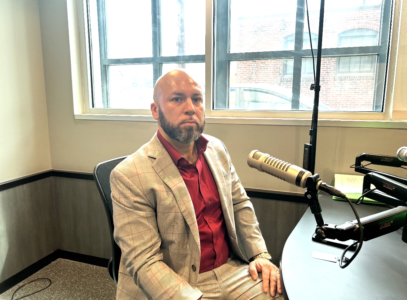 A man with a beard wearing a beige suit and red tie sitting in a studio with microphones and recording equipment.