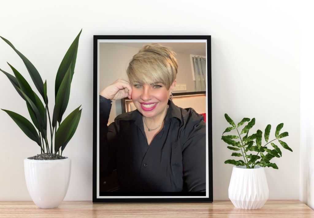 A smiling woman with short blonde hair wearing a black jacket and standing in front of a white wall.