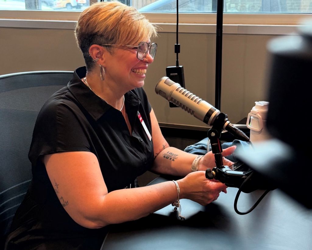 A smiling woman with short blonde hair wearing glasses and a black shirt, speaking into a microphone in a radio studio.