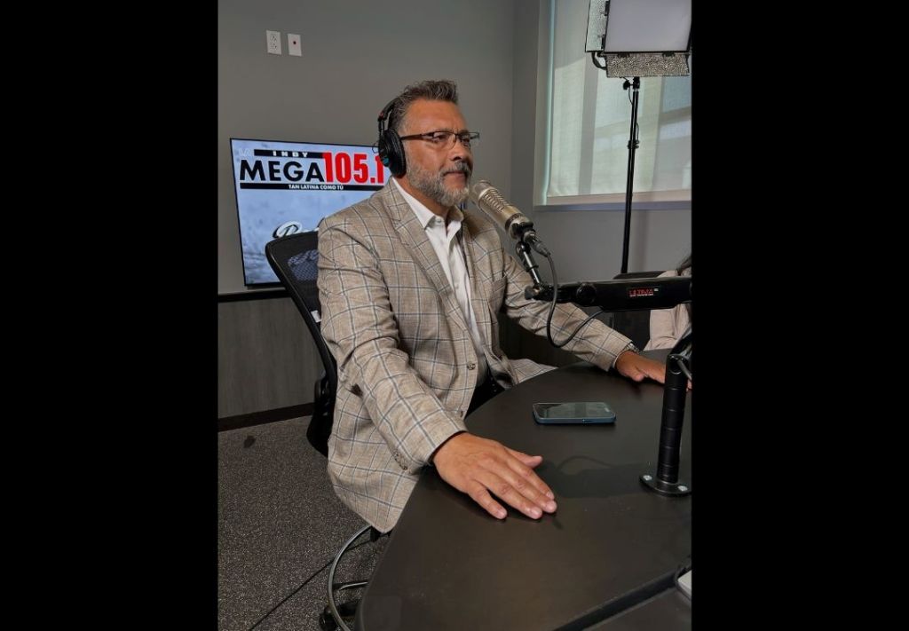 A man with a beard and glasses wearing a plaid suit jacket sits at a desk in a radio studio, with a "MEGA 105.1" logo visible on the screen behind him.