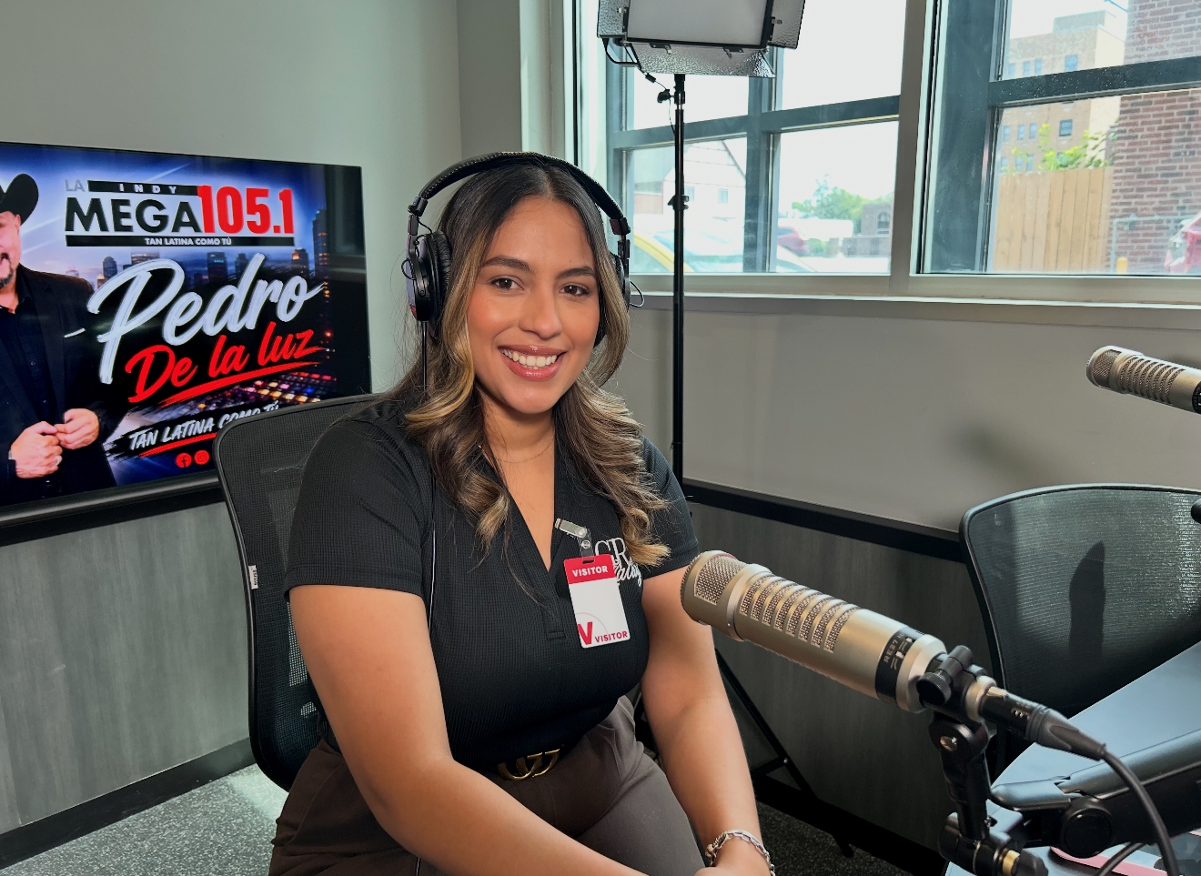 A smiling woman wearing headphones and a visitor badge sits in a radio studio, with a "Pedro de la Luz" logo visible on the screen behind her.