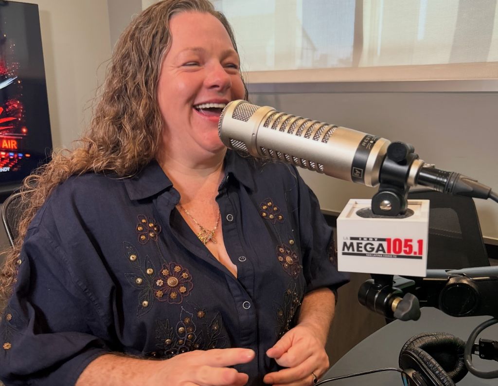 A smiling woman with curly hair speaking into a microphone with "MEGA 105.1" visible on the stand.