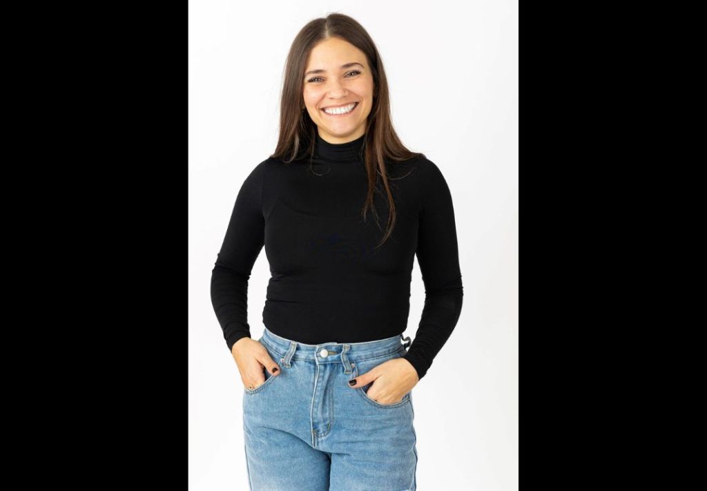 A smiling young woman with long dark hair wearing a black turtleneck and blue jeans, standing against a white background.