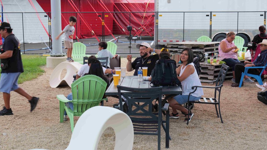 People sitting at tables outdoors, some wearing casual clothing, with a large red structure in the background.
