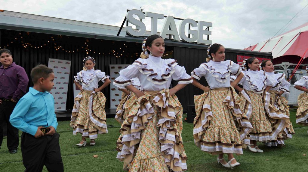 A group of young dancers in traditional Spanish-style dresses performing on a stage with a "STAGE" sign in the background.