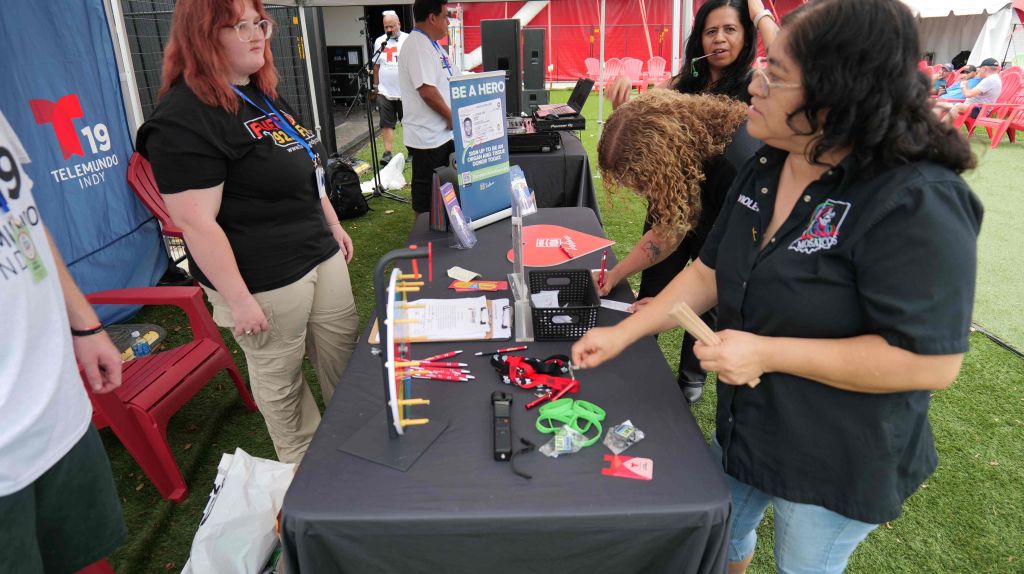 A group of people gathered at an event, with a table displaying various electronic and robotic devices. A sign on the table reads "Be a hero".