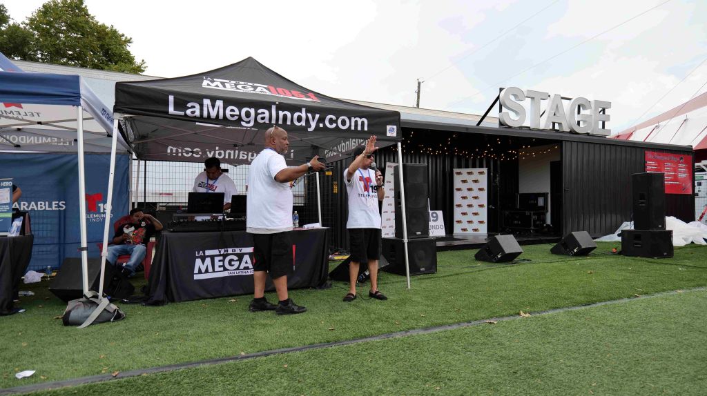 Outdoor event tent with "LaMegaIndy.com" branding, people working at the booth, and a large "MEGA" logo visible.