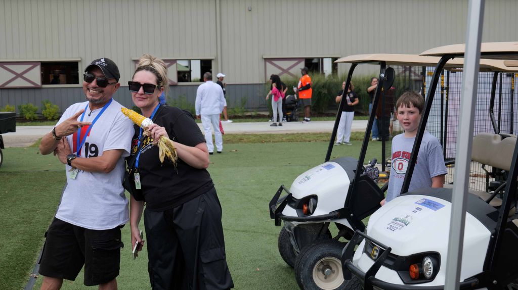 Two people wearing event staff uniforms with numbers, standing next to a golf cart with a helmet on it, in an outdoor setting with other people visible in the background.