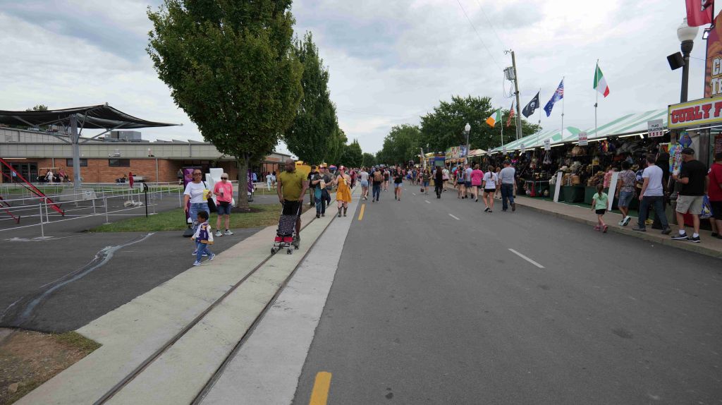 Crowded outdoor market with people walking, shopping, and riding scooters along a paved path lined with tents and stalls.