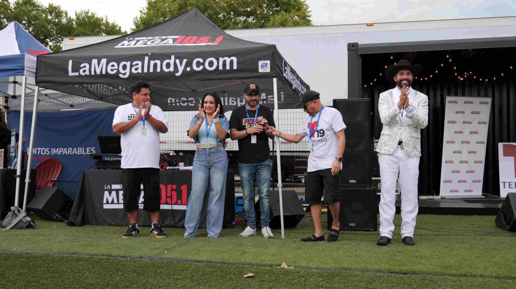 A group of people standing on a stage in front of a large tent with the "LaMegaIndy.com" logo, holding microphones and appearing to be at an event or broadcast.