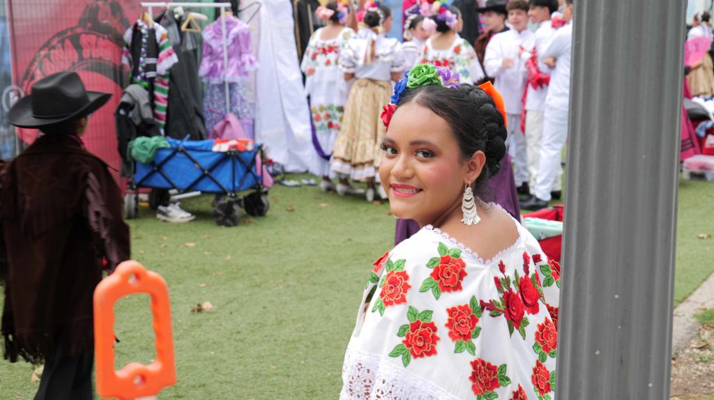 A smiling woman in traditional Mexican dress with floral embroidery, surrounded by a colorful crowd at what appears to be a festival or celebration.