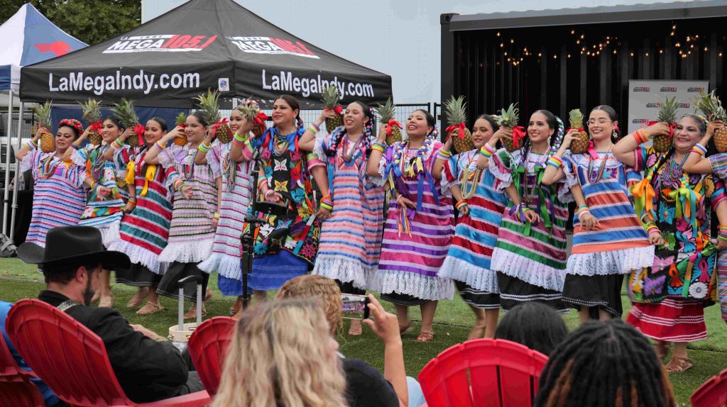 Colorfully dressed performers on stage at an outdoor event, with "LaMegaIndy.com" branding visible.