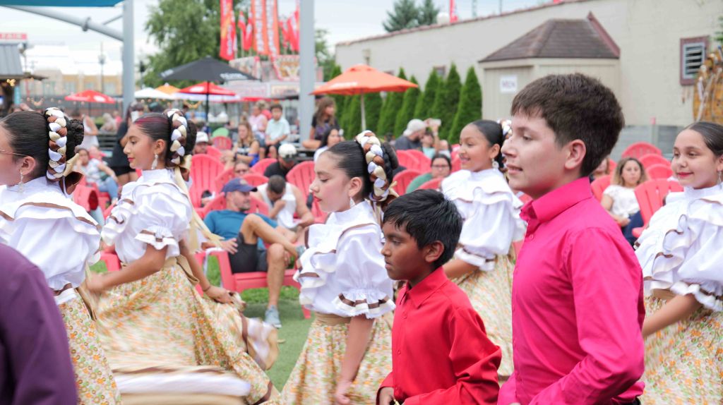 A crowd of people in traditional Latin American costumes performing at an outdoor festival, with red and white umbrellas and decorations visible in the background.