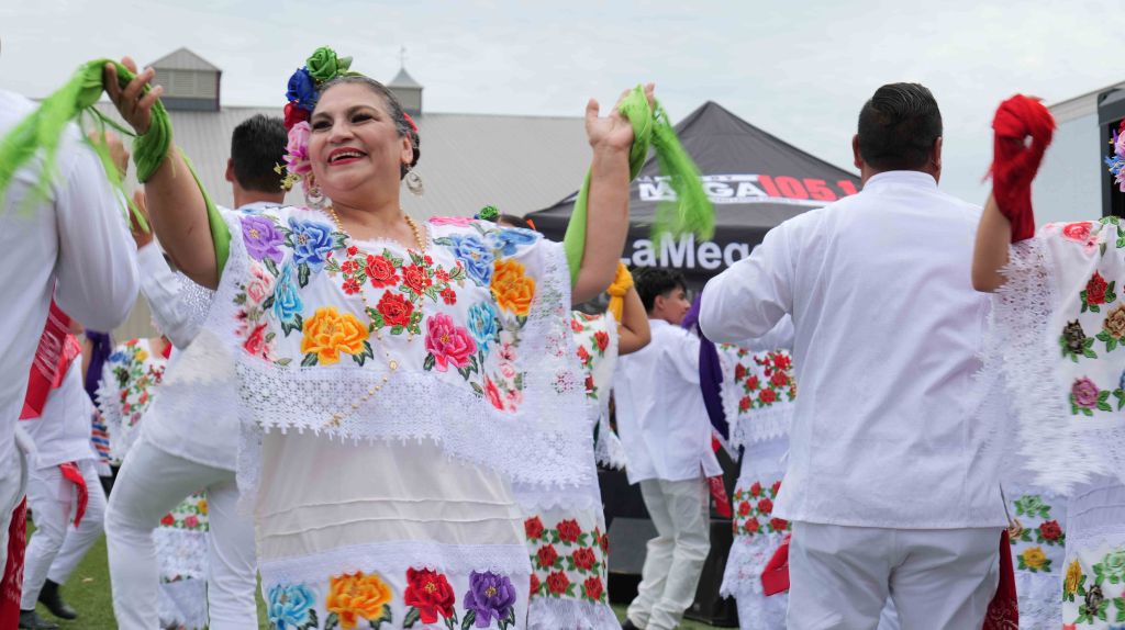 A smiling woman in a colorful, floral-patterned dress dances with others wearing traditional Mexican clothing at an outdoor event.
