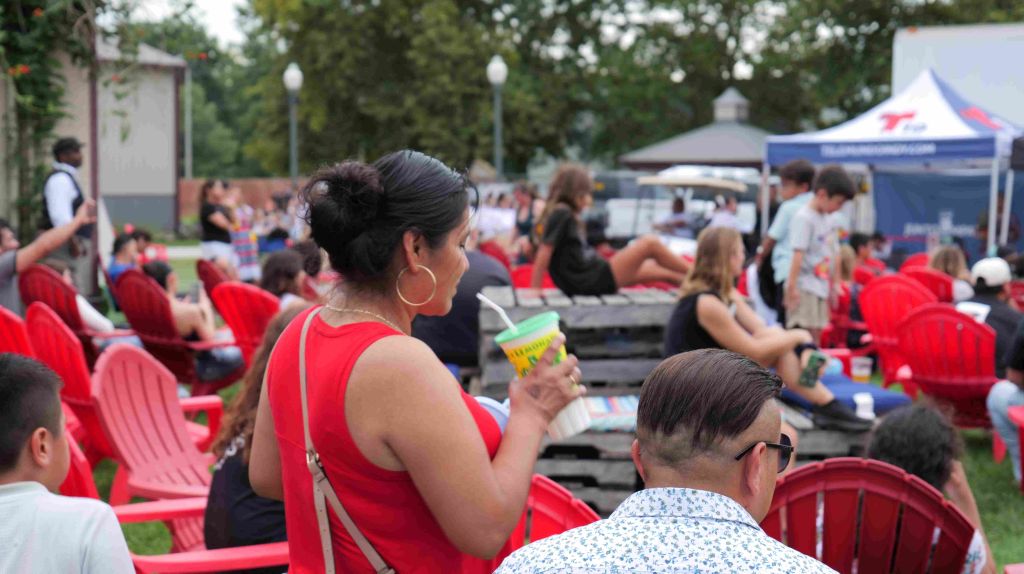 Outdoor market scene with people seated at red tables, enjoying food and drinks under trees.