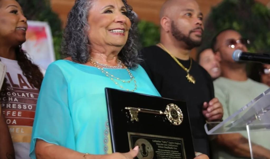A smiling older woman holding an award plaque, surrounded by a group of people, including a man with a beard.